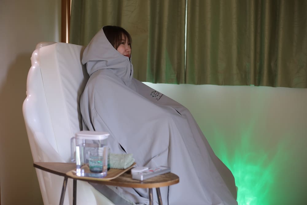 ルルオン専用ポンチョをまとい、緑のミストに包まれて全身を芯から温めるよもぎ蒸しの様子。サイドテーブルにはウェルカムドリンク